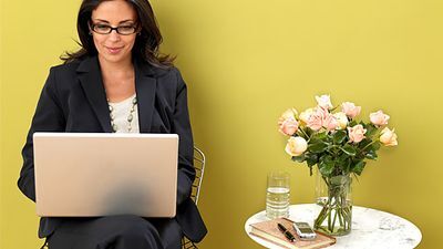 Woman using a laptop next to a marble top table with flowers, a cellphone, water, pen, and a notebook. 