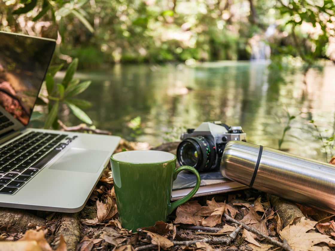 Photo by engin akyurt on Unsplash: Mug, thermos, Camera, and Laptop by a river