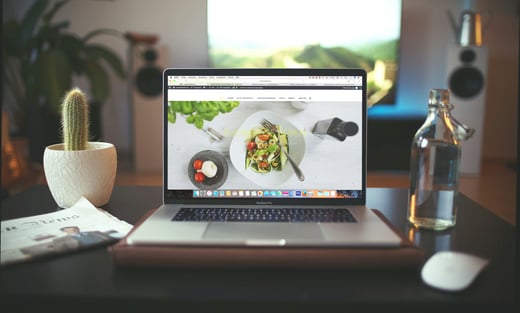 Photo by Igor Miske on Unsplash: Laptop on desk with cactus (left) and glass water bottle (right)..